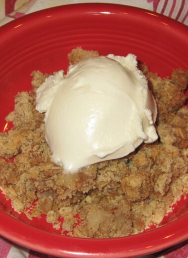 A red bowl with a scoop of gluten free apple crisp inside it and a scoop of vanilla ice cream on top.