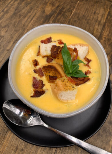A bowl full of gluten free beer cheese soup topped with croutons, crispy bacon and fresh basil.