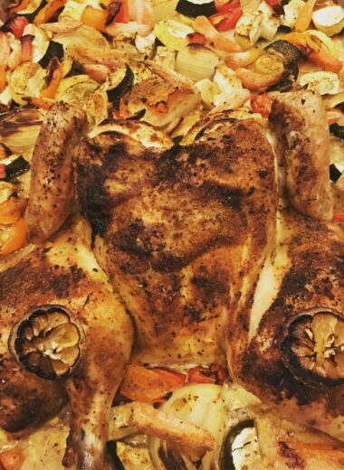 A sheet pan with a whole spatchcocked roasted chicken and vegetables and lemons on it.