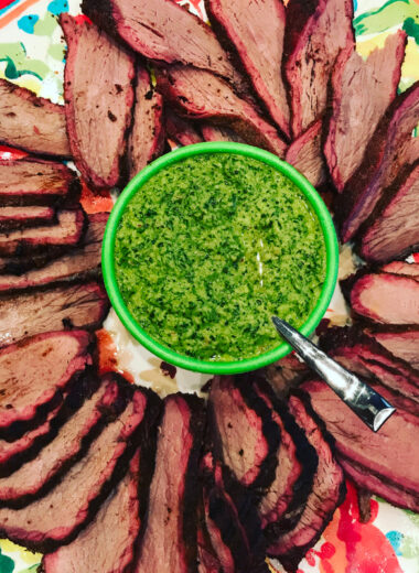 A colorful platter with smoked meat in a circle around a bowl full of bright green chimichurri sauce.