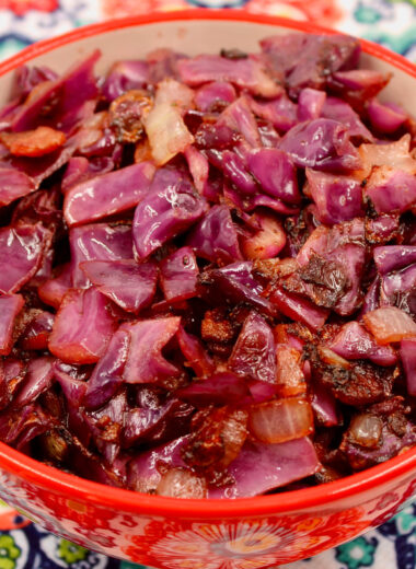A red bowl sitting on top of a colorful mat holding cooked cabbage and bacon.