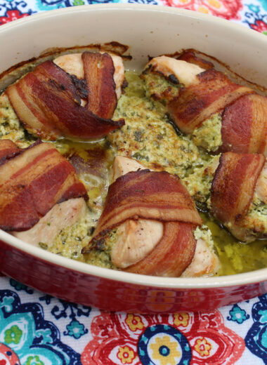 A baking dish sits on a multicolored background with chicken wrapped in bacon and a creamy filling inside of it.