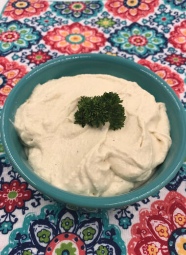 A colorful mat with a blue bowl filled with mashed cauliflower in it and a sprig of parsley on top.