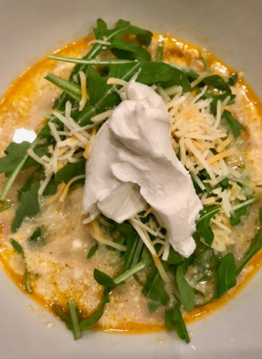 A white bowl filled with creamy soup topped with arugula, shredded cheese and sour cream.