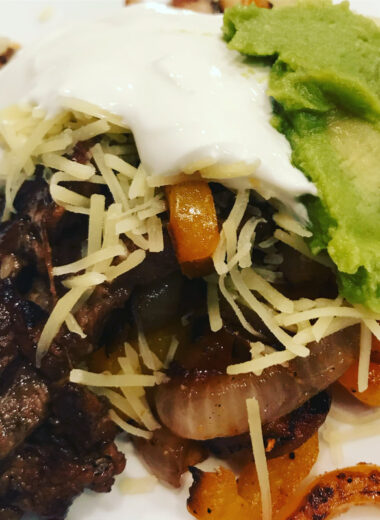 A white plate with grilled onions, peppers and steak on it topped with shredded cheese, guacamole and sour cream.