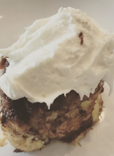 A cinnamon bun on a white plate with frosting on top of the bun.