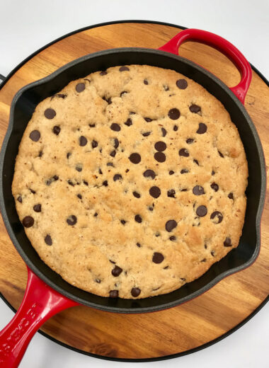 A wooden board is under a cast iron skillet with chocolate chip cookie baked inside of it.