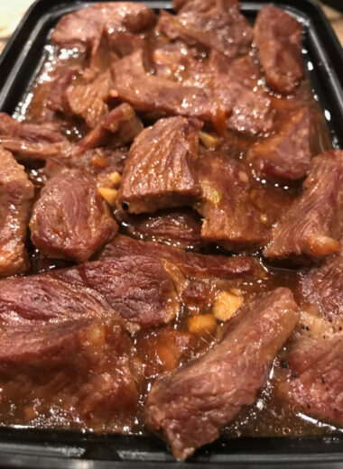 A black container holding cooked beef pieces with brown sauce on it.