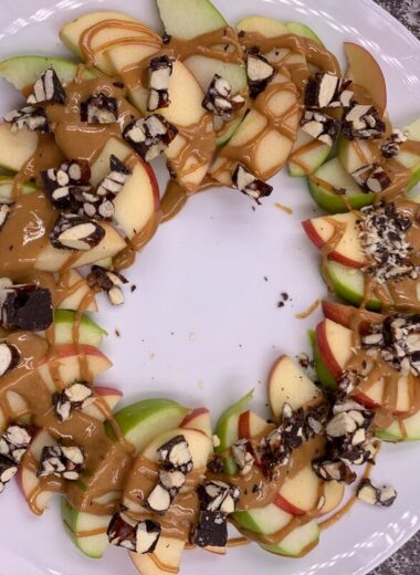 A white plate with apple slices in a circle with nuts, chocolate and peanut butter on top.