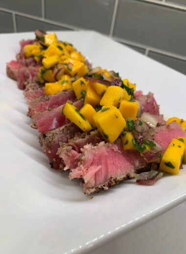 A white plate with seared rare tuna sliced on it topped with mangoes and onions.