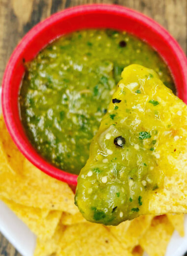 A red bowl filled with bright green tomatillo salsa and tortilla chips on the side to dip in it. One chip has salsa on it.