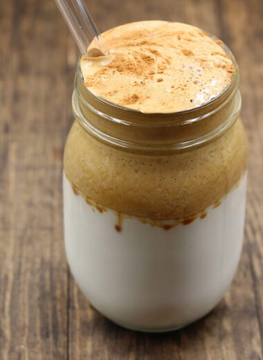 A clear glass of milk on the bottom and foamy coffee on the top with a straw in it sitting on a wooden counter.