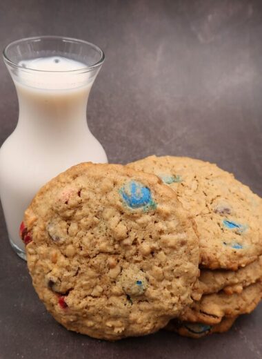 Peanut Butter Oatmeal Chocolate Chip MnM cookies stacked with one leaning against them and a cute jar of milk beside them.
