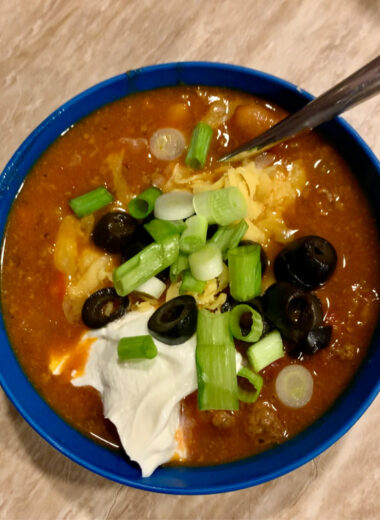 A blue bowl full of chili with green onion slices, black olives and sour cream on top.