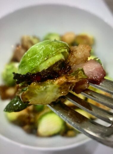 A fork holding crispy Brussels sprouts with bacon over a white bowl with more crispy Brussels Sprouts in it.